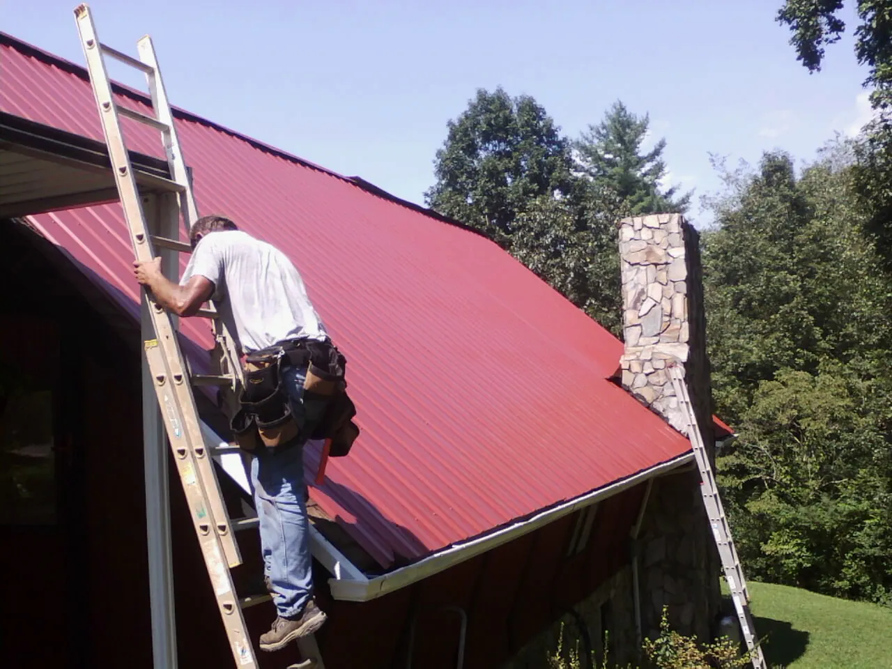 Roofer on a ladder beside a red metal roof and stone chimney — Soffit & Fascia Repair by Roofing Brothers in Grizzly Flats